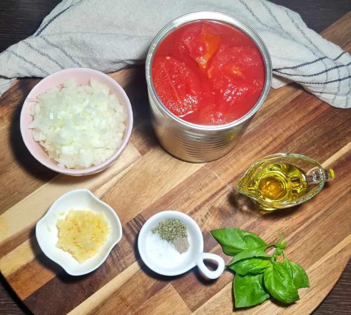 Ingredients for homemade marinara sauce including canned tomatoes, chopped onions, minced garlic, olive oil, basil leaves, and a small bowl of salt, pepper, and oregano on a wooden cutting board.