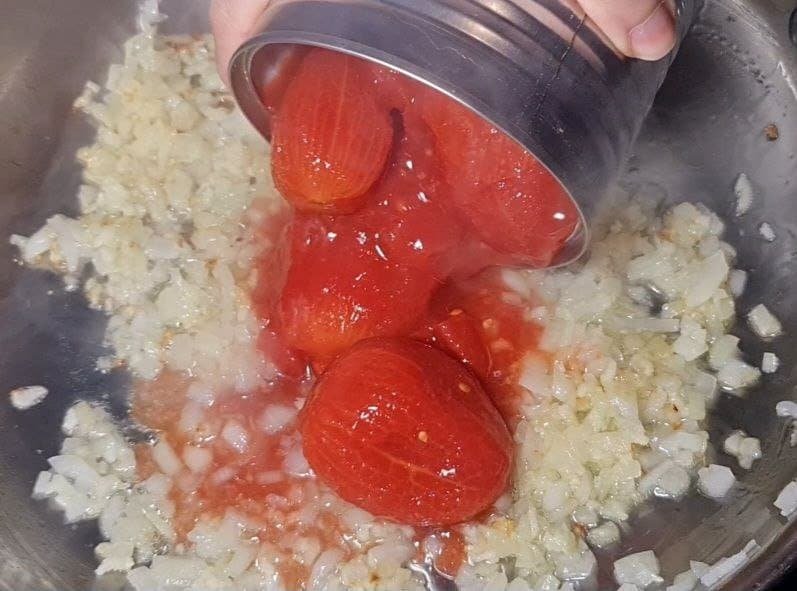 Pouring canned whole tomatoes into a pan with sautéed onions for homemade marinara sauce.