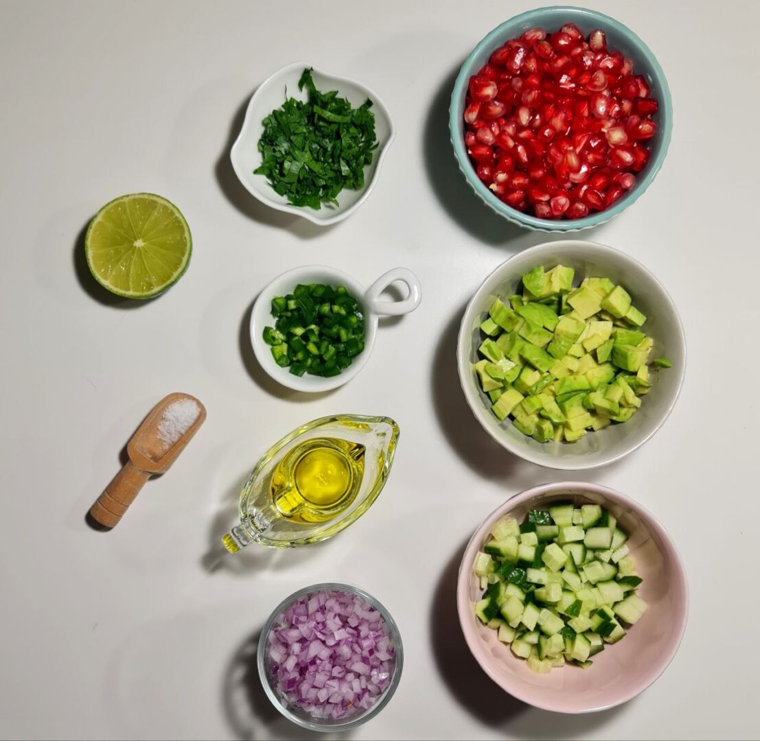 A collection of ingredients including pomegranate seeds, diced avocado, diced cucumber, chopped green herbs, chopped green chili peppers, diced red onions, olive oil, salt, and half a lime, arranged on a white surface. Fresh ingredients for making vibrant and delicious pomegranate salsa.