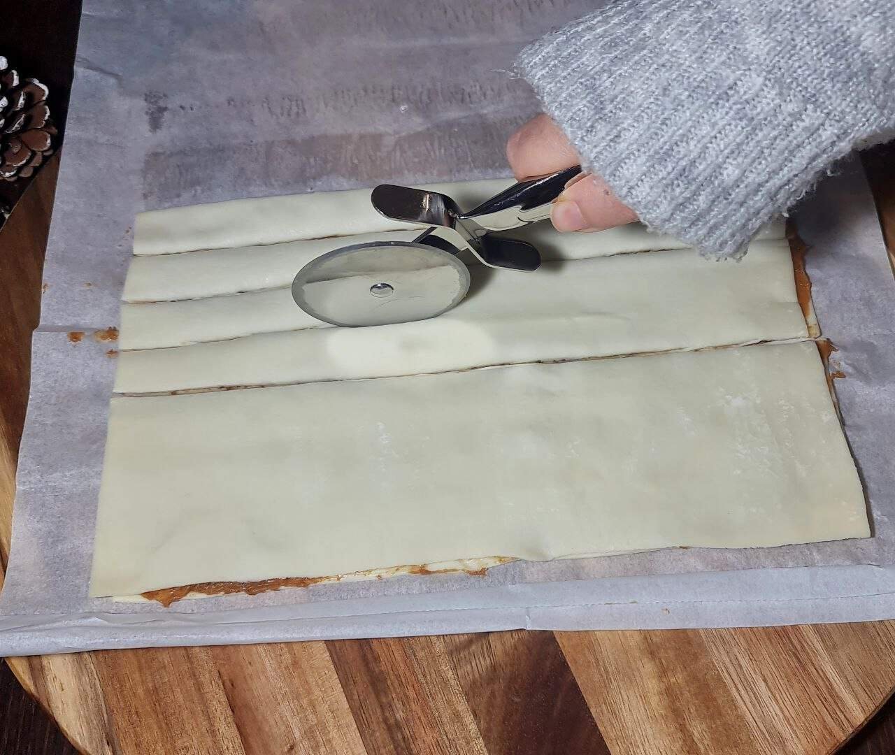 A hand holding a pizza cutter, cutting strips of dough on a piece of parchment paper. 