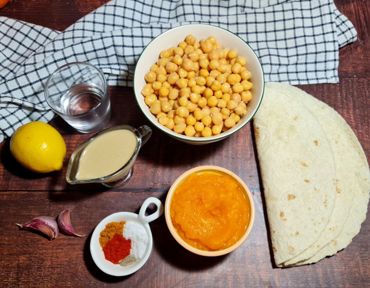 Ingredients for pumpkin hummus on a wooden table, including chickpeas, tahini, lemon, spices, garlic, water, and tortillas. Gather all the ingredients for the perfect pumpkin hummus.