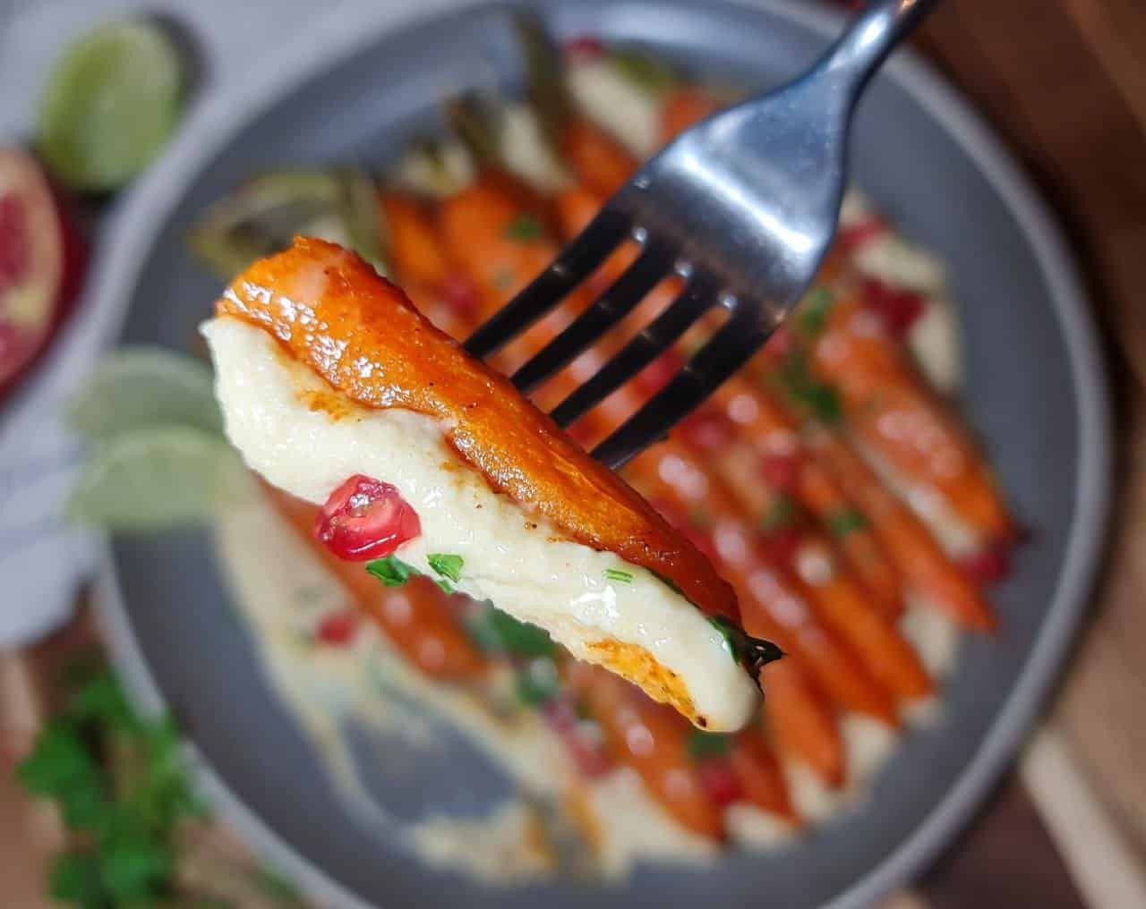  A fork holding a roasted carrot dipped in creamy hummus, garnished with pomegranate and parsley. Close-up of a roasted carrot dipped in hummus.