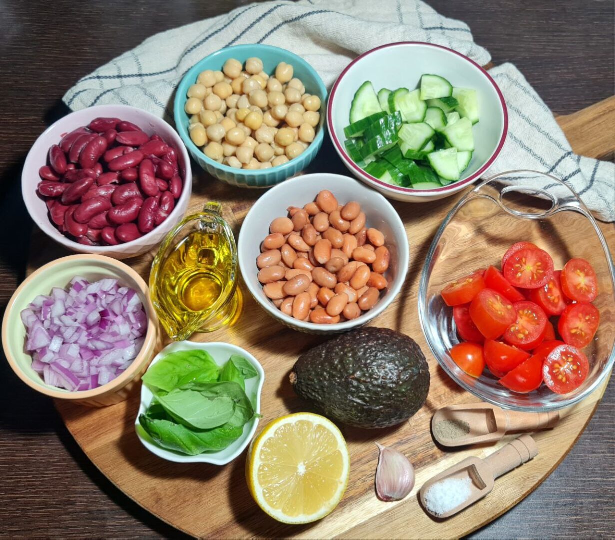 Fresh ingredients for three bean salad including kidney beans, black beans, chickpeas, chopped red onions, cherry tomatoes, avocado, cucumber, and basil leaves.