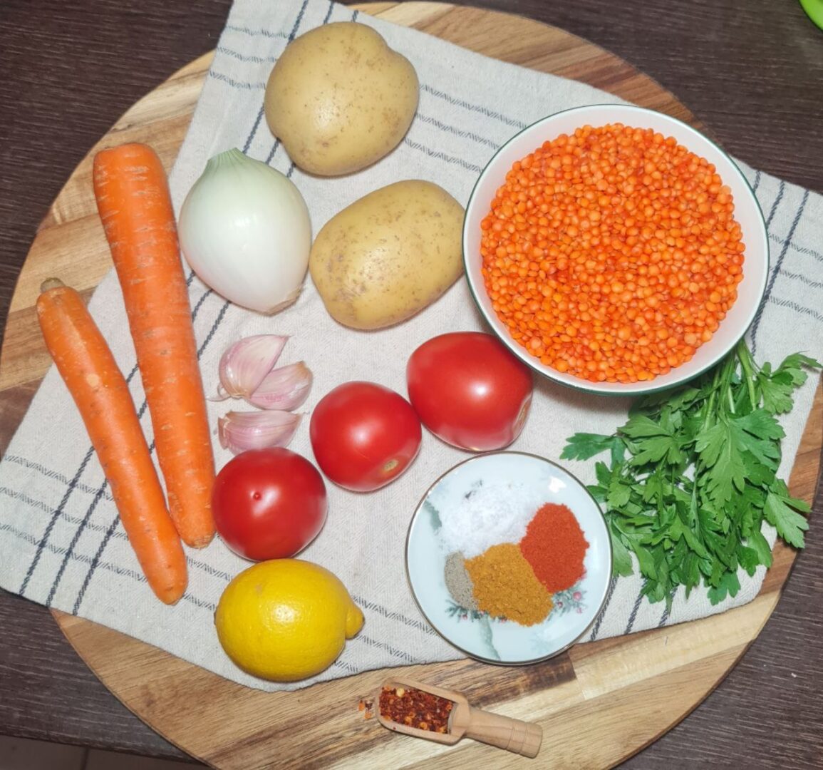 Fresh vegetables and spices arranged on a wooden board, including carrots, onion, potatoes, tomatoes, garlic, lemon, red lentils, and various spices. Fresh ingredients and spices ready to be transformed into delicious Turkish lentil soup.