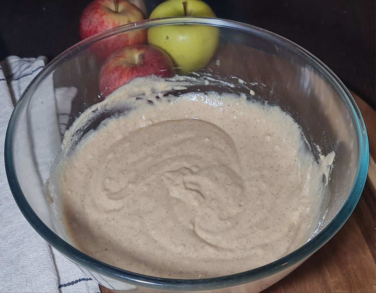  A glass bowl with a light brown batter mixture, three apples (two red and one green) on a wooden surface, and a white cloth with a blue stripe pattern partially visible. Mixing the batter for vegan apple cake with fresh apples and simple ingredients.