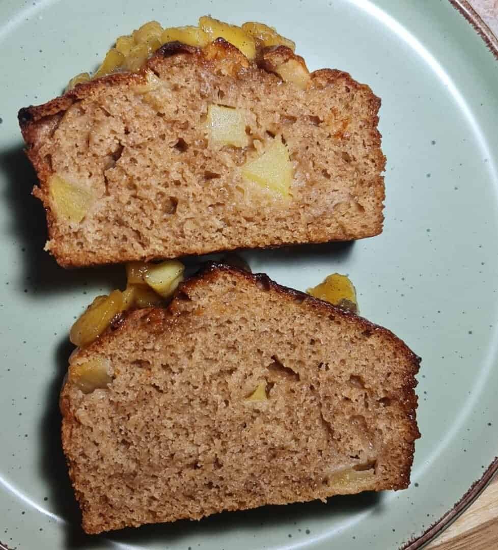 A loaf of vegan apple cake with visible apple chunks, sliced on a green plate, surrounded by whole red and green apples. 