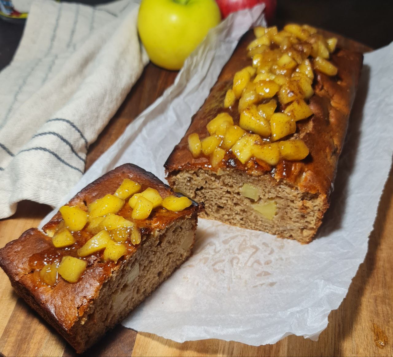 A loaf of vegan apple cake with visible apple chunks, sliced on a green plate, surrounded by whole red and green apples. Moist and delicious vegan apple cake with fresh apple chunks, perfect for a homemade treat.