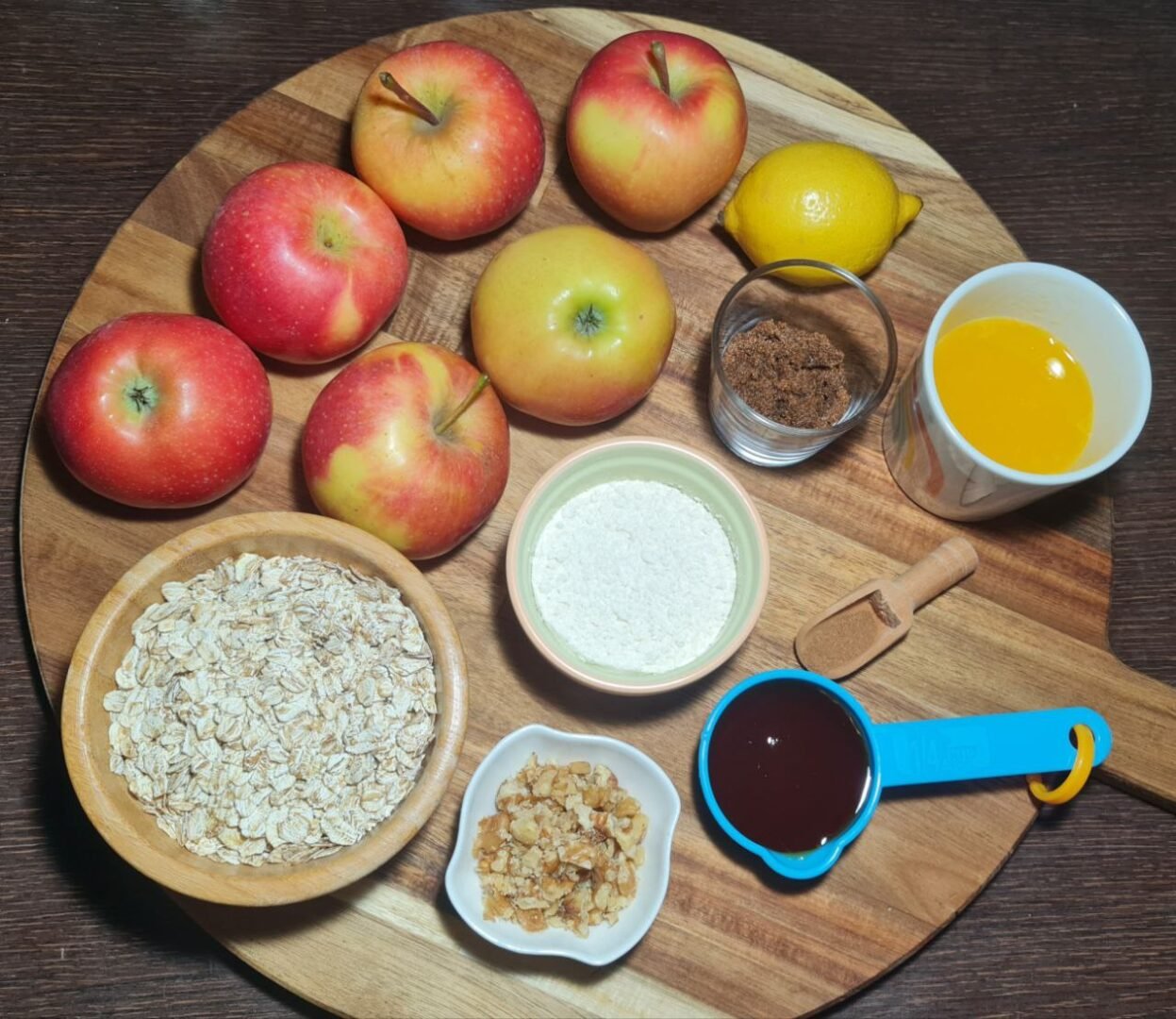 Ingredients for vegan apple crisp arranged on a wooden board, including apples, lemon, brown sugar, melted butter, flour, rolled oats, chopped walnuts, syrup, and cinnamon. Fresh ingredients for a delicious vegan apple crisp.