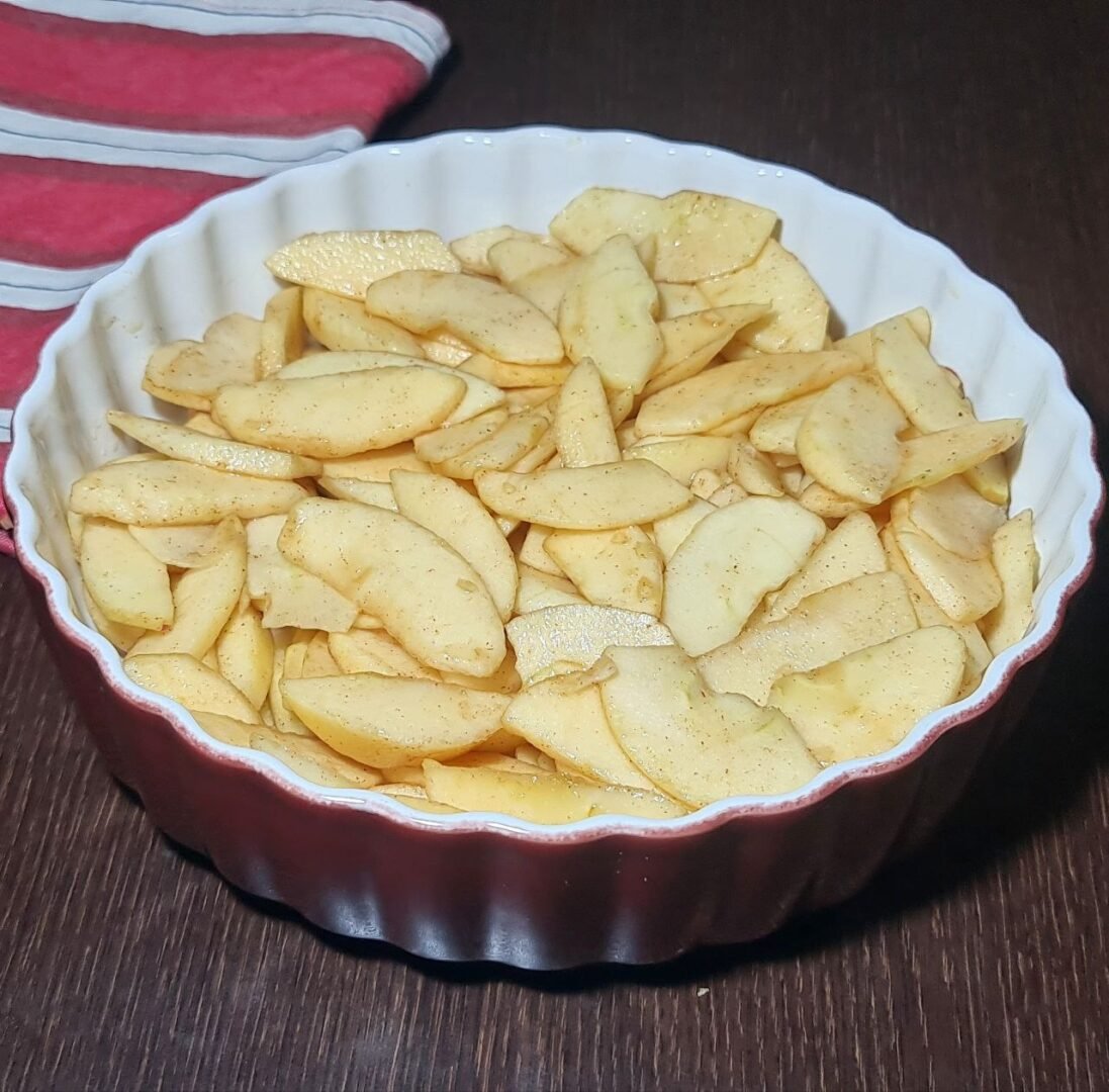 Sliced apples seasoned with cinnamon in a white and red baking dish, ready for making vegan apple crisp. Sliced apples seasoned with cinnamon in a baking dish, prepared for a vegan apple crisp.