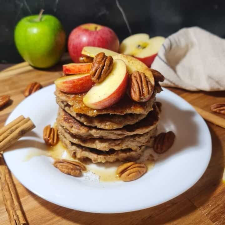 A stack of vegan pancakes topped with apple slices, pecans, and syrup being poured over them.