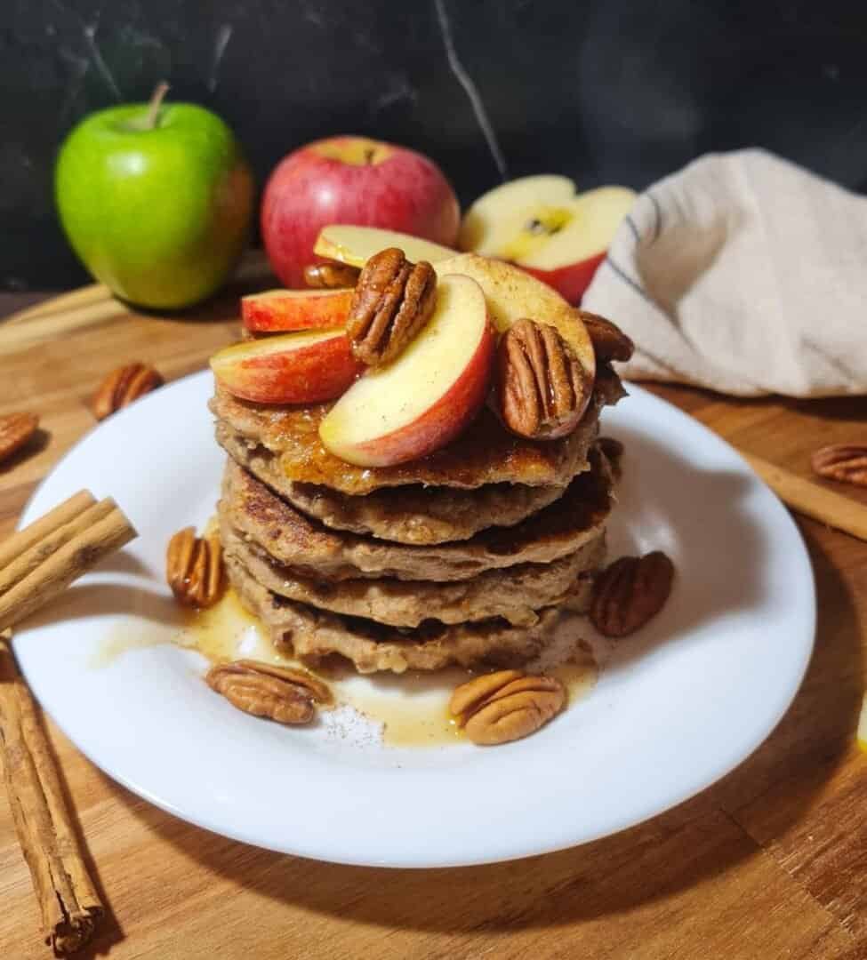 A stack of vegan pancakes topped with apple slices, pecans, and syrup being poured over them.