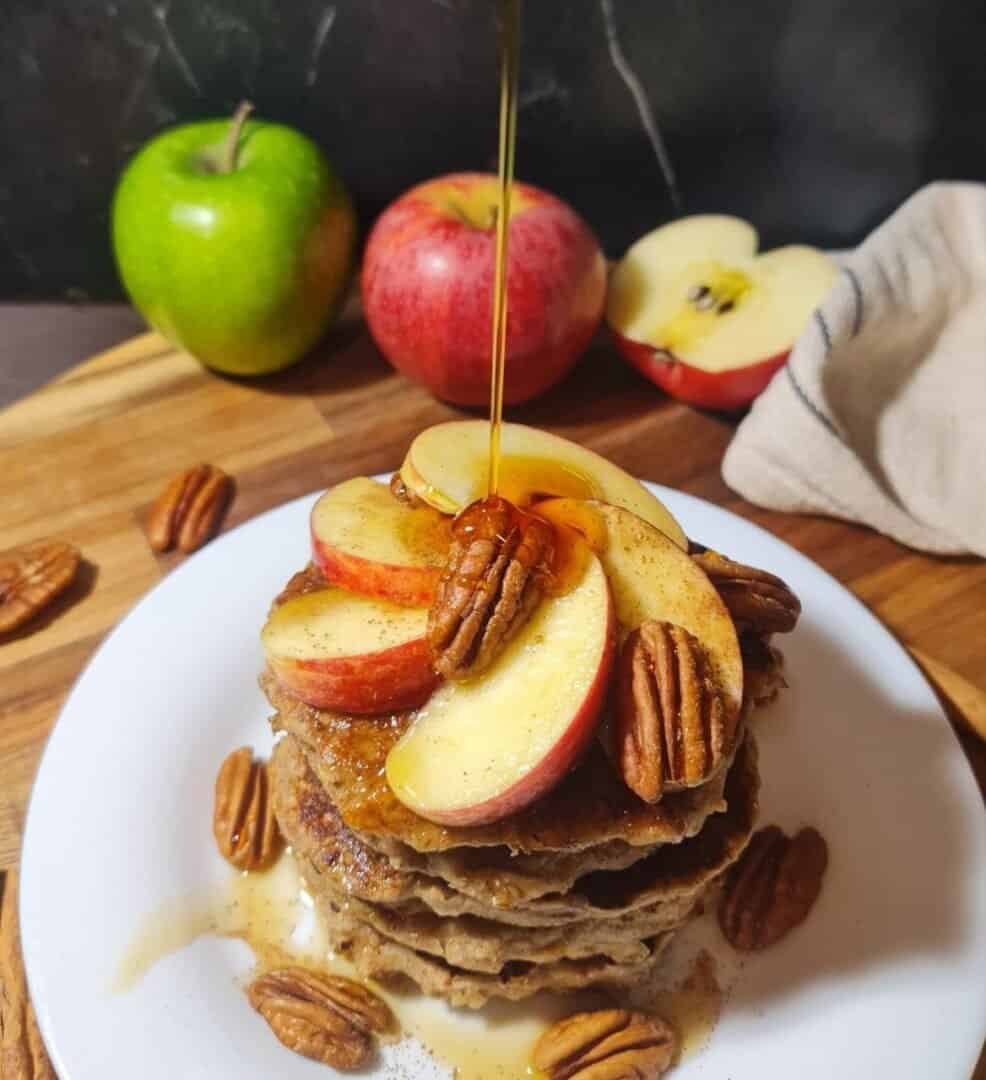 A stack of vegan apple pecan pancakes topped with fresh apple slices and pecans, drizzled with syrup.