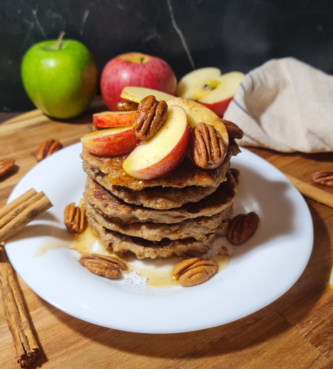 A stack of vegan pancakes topped with apple slices, pecans, and syrup being poured over them.