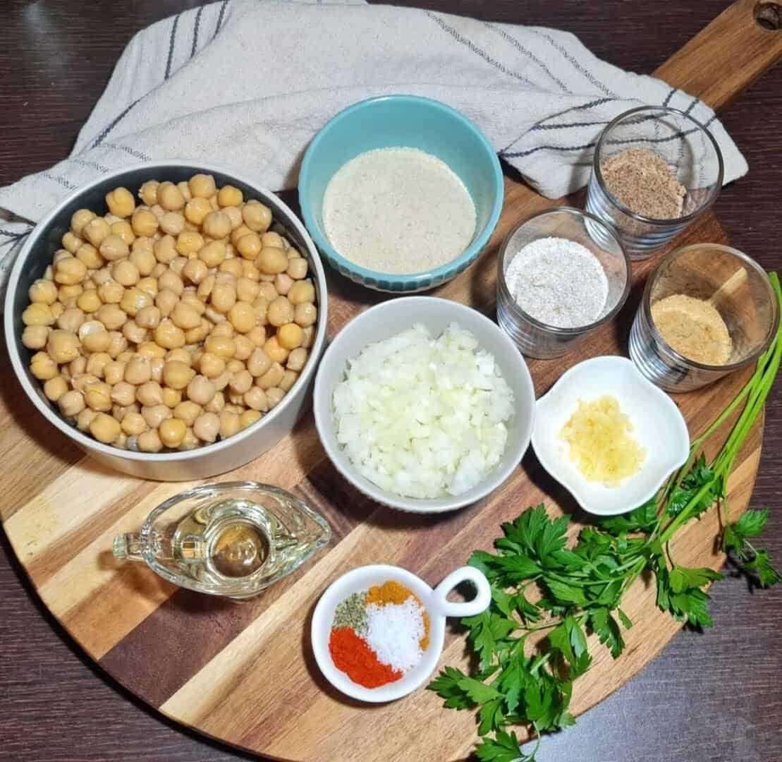  Ingredients for vegan chickpea meatballs including chickpeas, breadcrumbs, chopped onions, minced garlic, spices, and fresh parsley arranged on a wooden board.