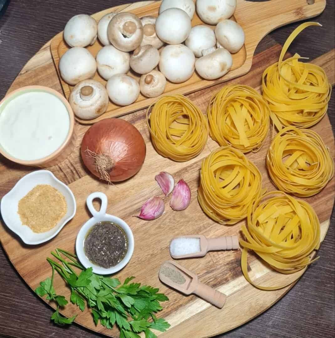 Ingredients for vegan creamy mushroom and truffle pasta on a wooden board, including mushrooms, fettuccine pasta nests, onion, garlic, parsley, truffle paste, vegan cream, nutritional yeast, salt, and pepper. Fresh ingredients for a delicious vegan creamy mushroom and truffle pasta.