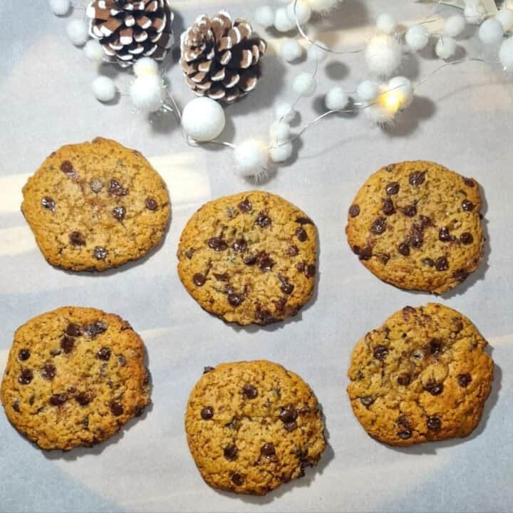 Six vegan gingerbread chocolate chip cookies on parchment paper with festive pinecones and white decorative lights in the background.