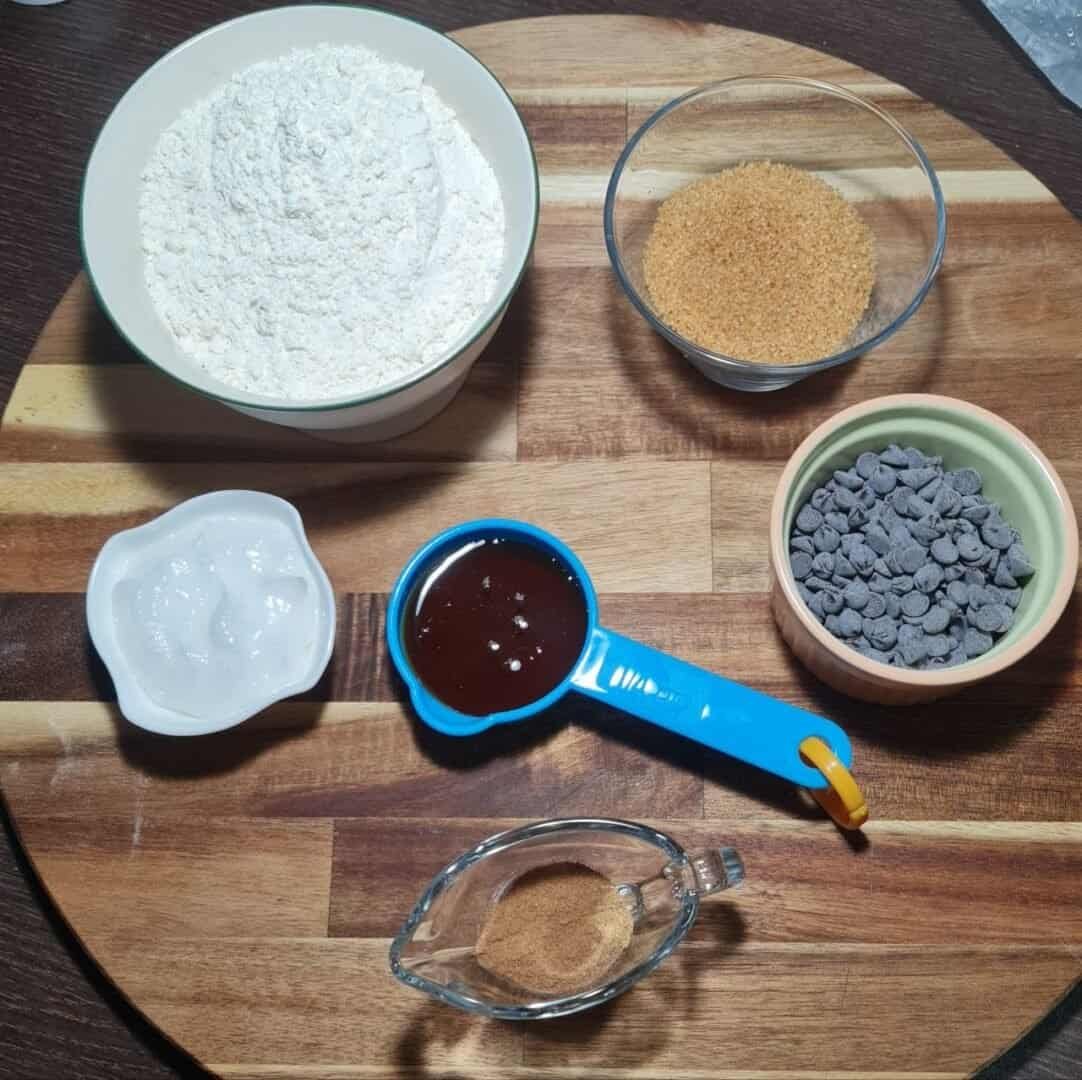 Ingredients for vegan gingerbread chocolate chip cookies arranged on a wooden board, including flour, brown sugar, chocolate chips, coconut oil, molasses, and ground ginger.