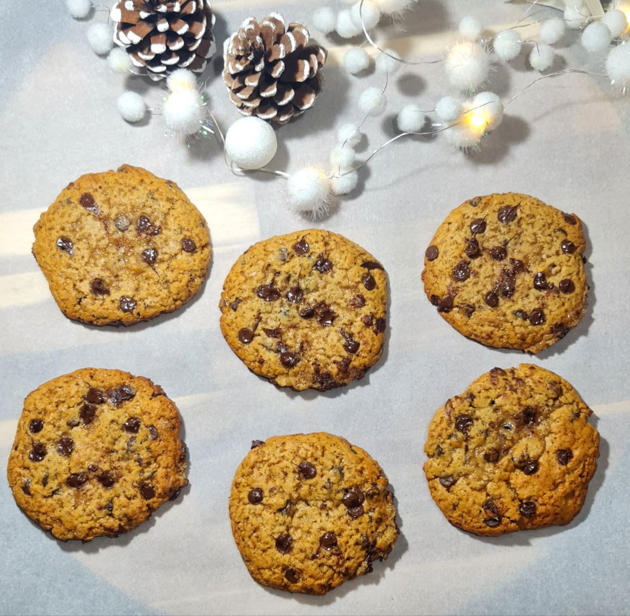 Six vegan gingerbread chocolate chip cookies on parchment paper with festive pinecones and white decorative lights in the background.