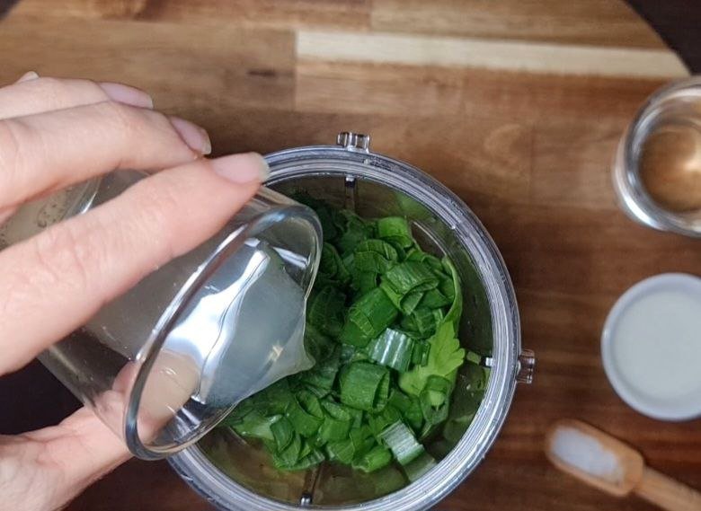  Pouring water over green onions and herbs in a blender for vegan green goddess dressing. Blending fresh herbs and greens to create a vibrant green goddess dressing.