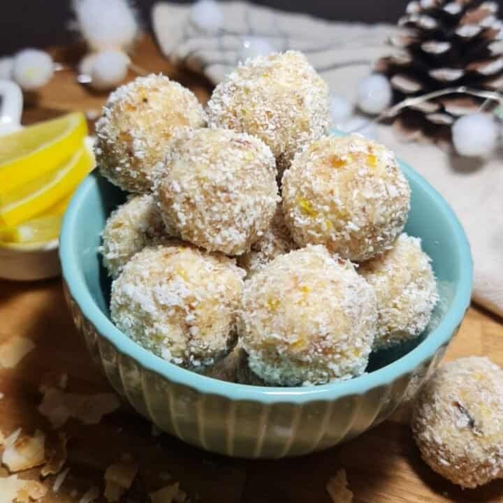 A bowl filled with vegan lemon coconut balls, surrounded by pine cones and lemon slices on a wooden surface.