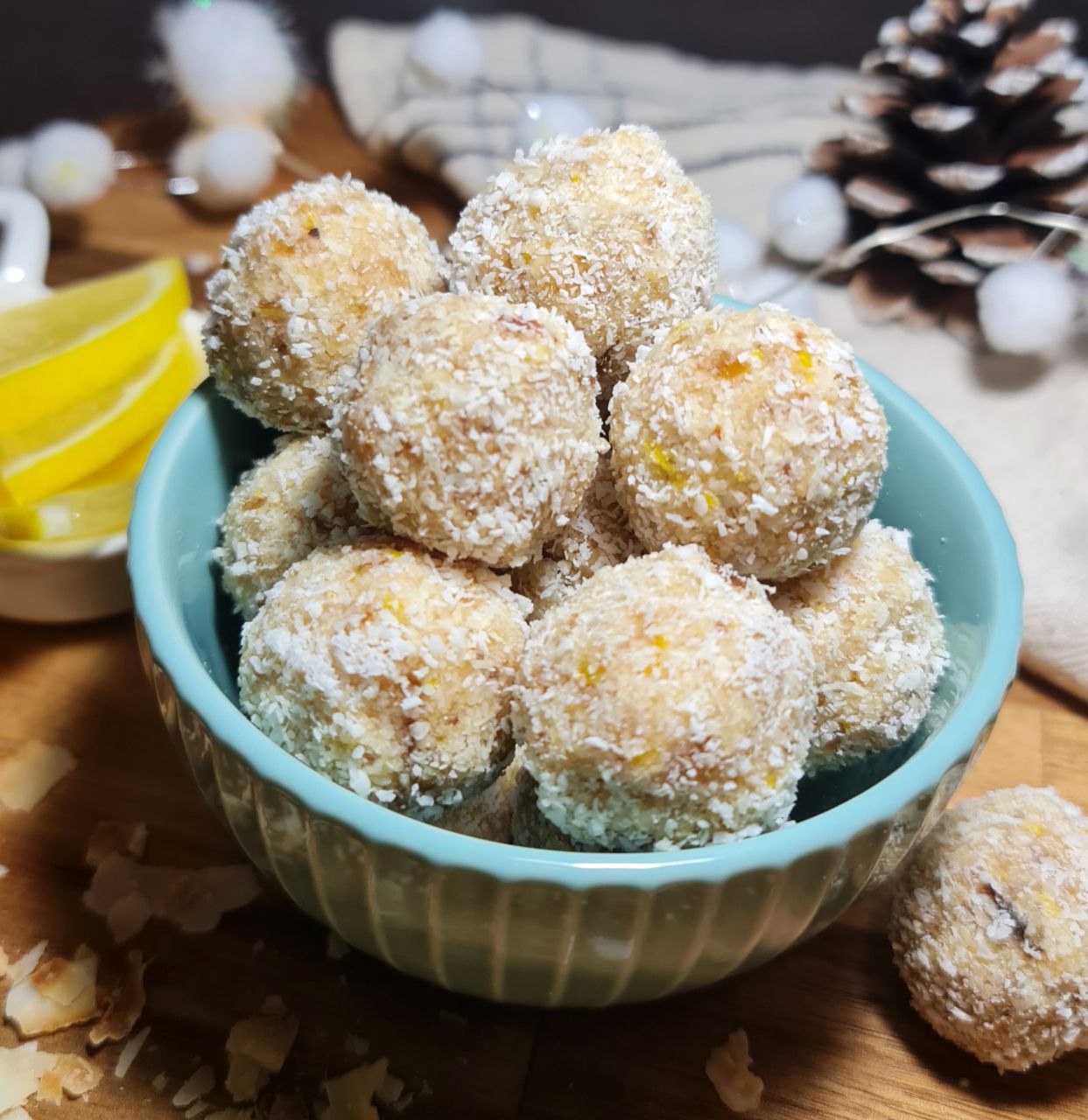 A bowl filled with vegan lemon coconut balls, surrounded by pine cones and lemon slices on a wooden surface.