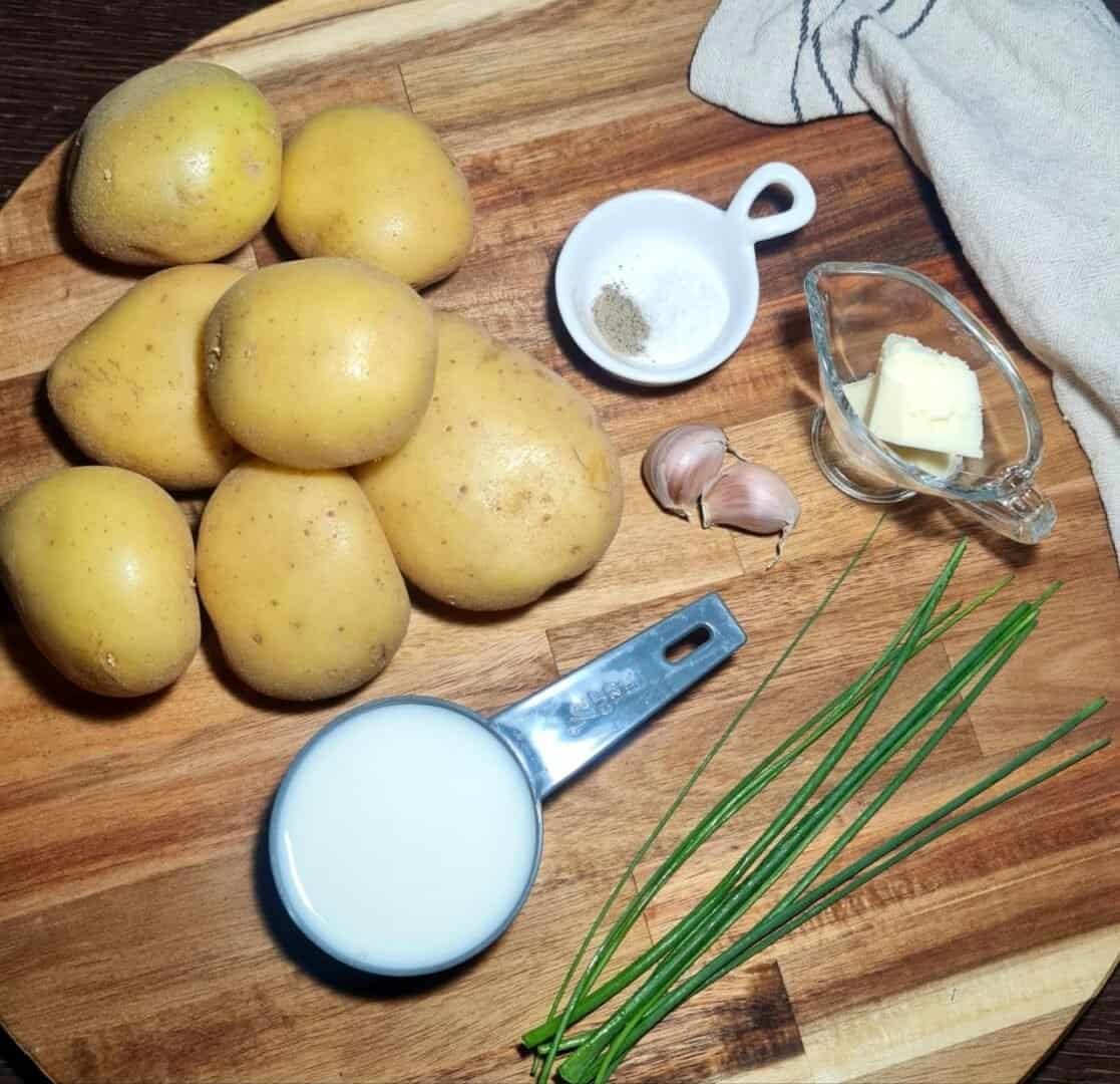  Ingredients for vegan mashed potatoes recipe including yellow potatoes, garlic, milk, butter, salt, pepper, and fresh chives arranged on a wooden cutting board. Fresh and wholesome ingredients for making delicious vegan mashed potatoes.