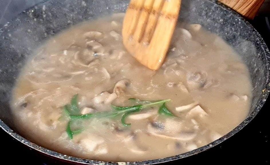 Cooking vegan mushroom gravy in a pan with a wooden spatula stirring the mixture. Stirring vegan mushroom gravy in a pan with fresh herbs.