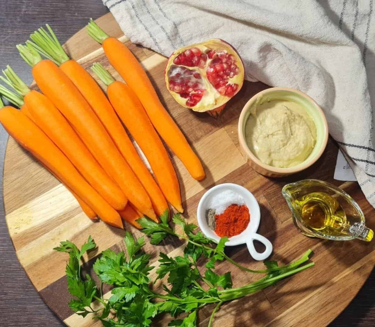 Fresh carrots, hummus, pomegranate, parsley, and spices arranged on a wooden board.  Ingredients for whole roasted carrots with hummus.