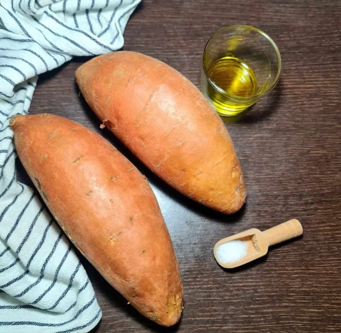 Two raw sweet potatoes, a glass of olive oil, and a small scoop of salt on a wooden surface with a striped cloth.