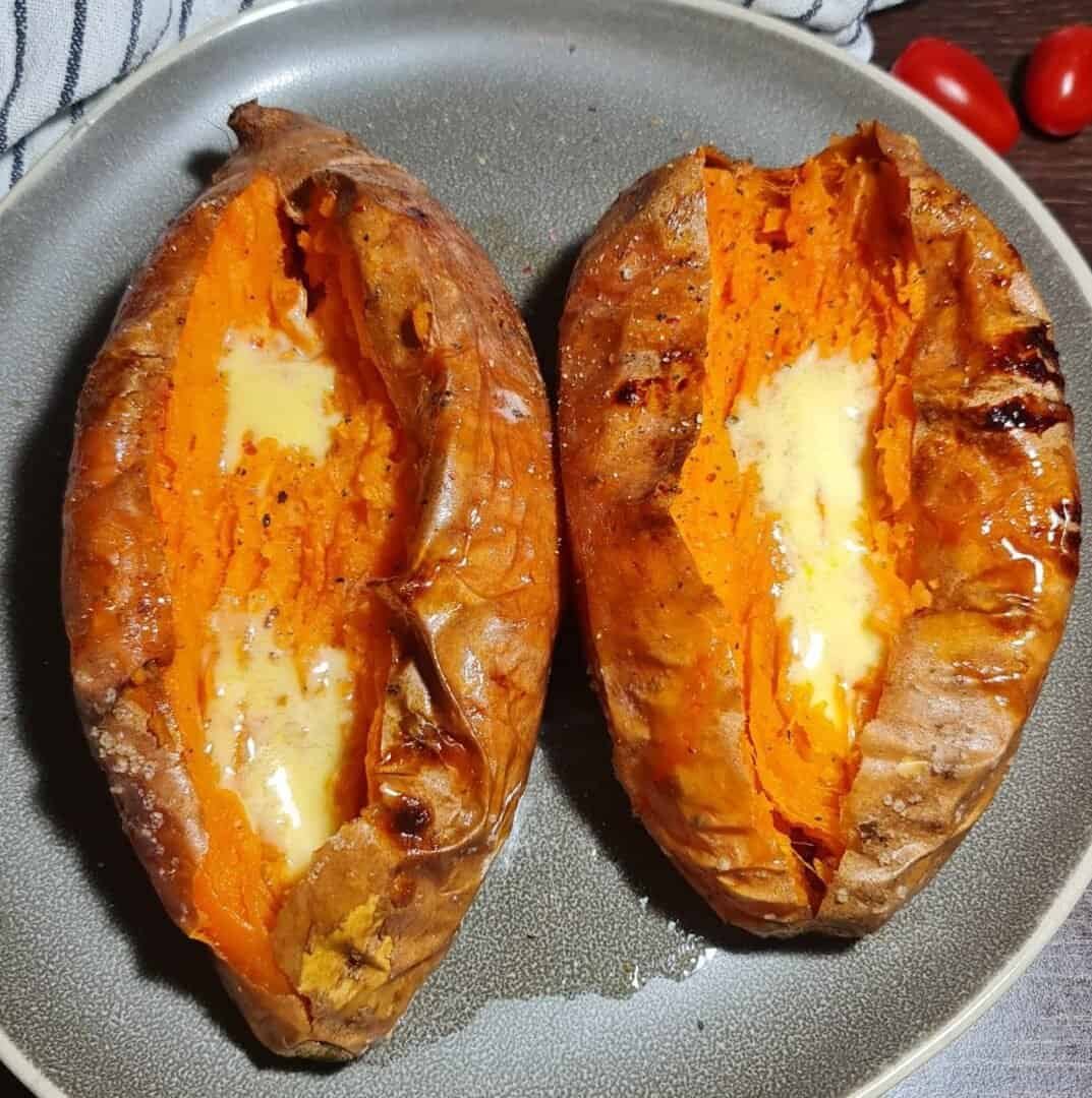 Two baked sweet potatoes with melted butter on a plate, garnished with cherry tomatoes and a striped cloth napkin.