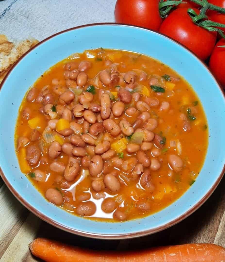 A bowl of pinto bean soup garnished with herbs, surrounded by fresh ingredients like tomatoes and carrots.