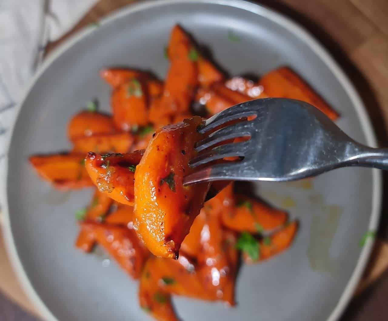  Close-up of a fork holding a piece of glazed brown sugar carrot with a plate of more glazed carrots in the background.