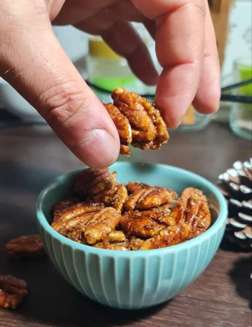 A hand holding candied pecans above a bowl filled with more candied pecans.