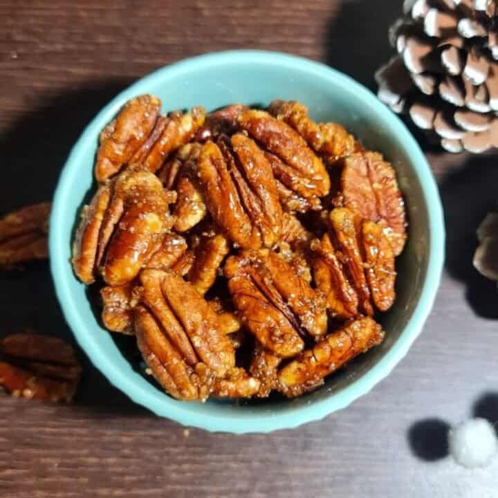 A bowl of candied pecans on a wooden table with a pine cone in the background.