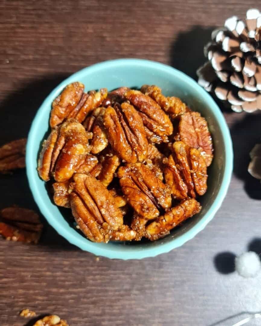 A bowl of candied pecans on a wooden table with a pine cone in the background.