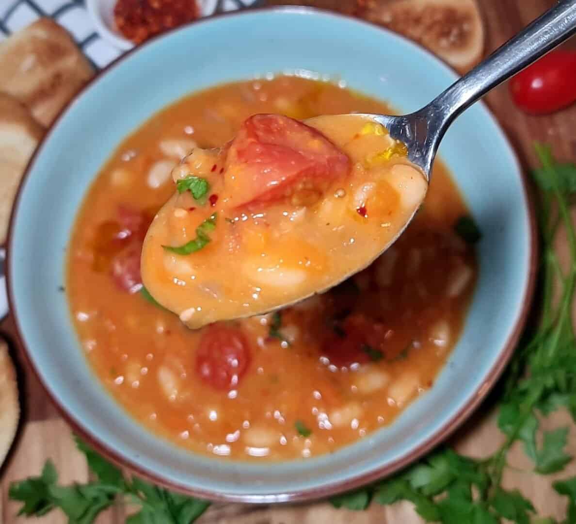 A close-up of a spoonful of cannellini bean soup with tomatoes and herbs, held above a bowl of the same soup.