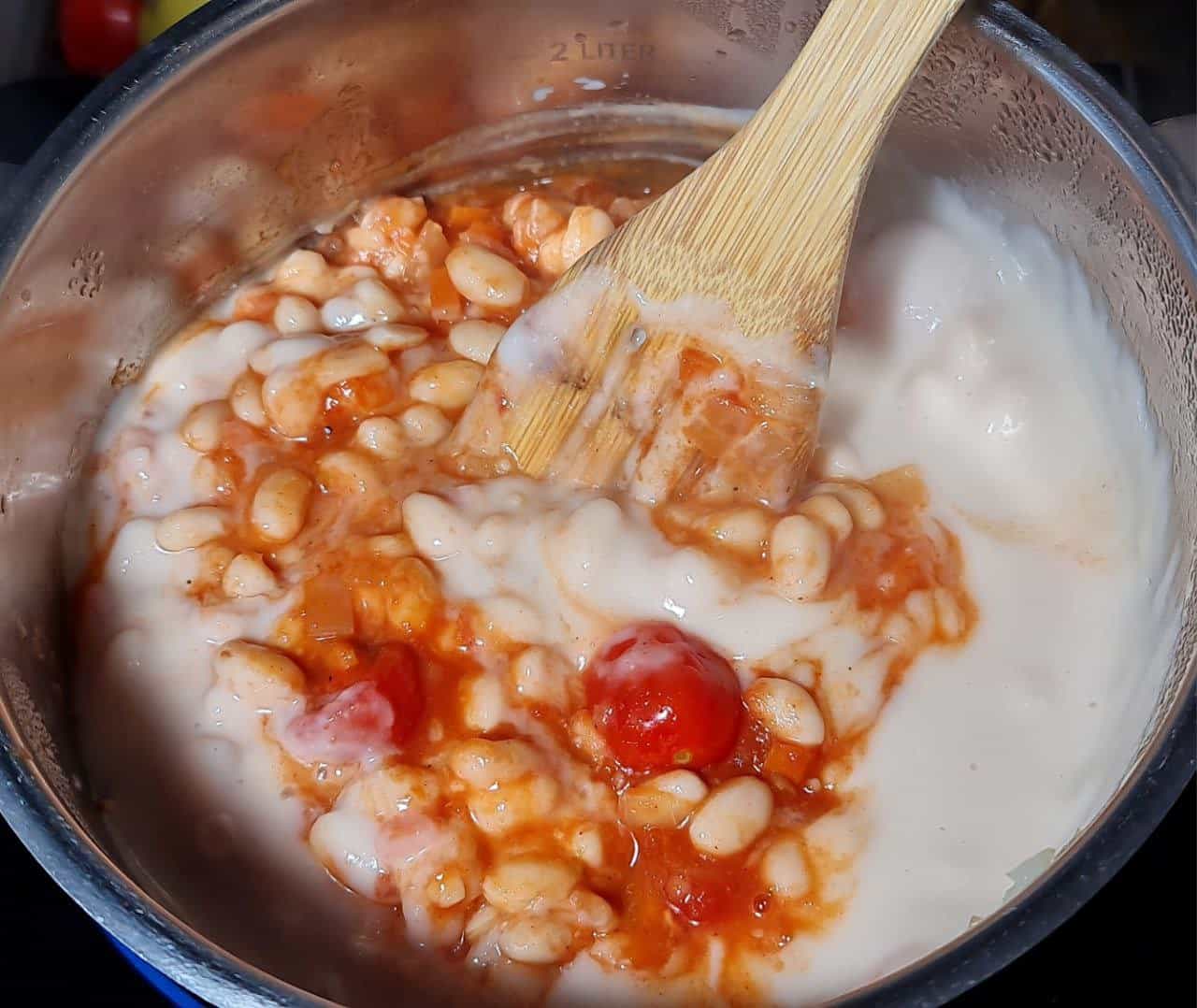 A pot of creamy cannellini bean soup being stirred with a wooden spoon, showing beans, tomatoes, and a rich broth.