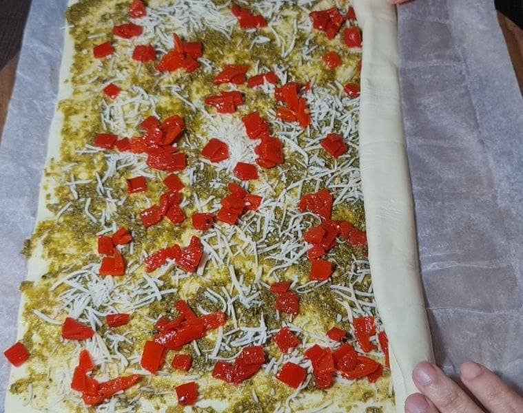 Preparing Christmas pinwheel appetizer with rolled dough, pesto, cheese, and red bell peppers.