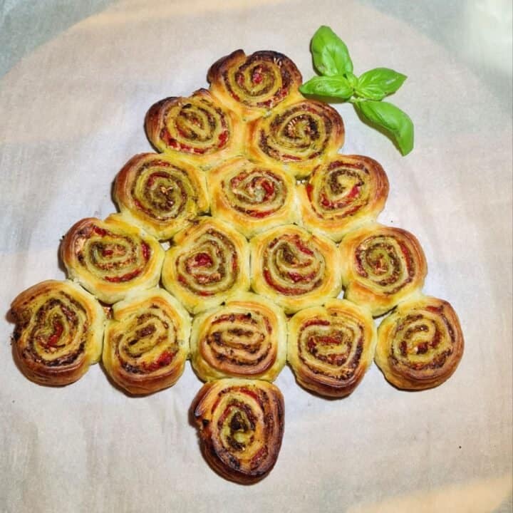 Christmas tree-shaped pinwheel appetizer made with puff pastry, pesto, and red bell pepper, garnished with fresh basil leaves.