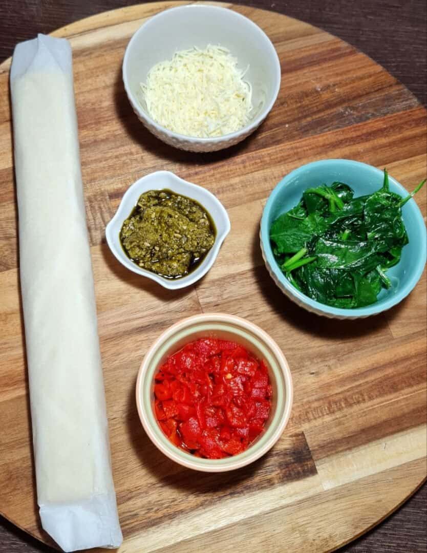  Ingredients for Christmas pinwheel appetizer including rolled dough, pesto, cheese, and diced red bell peppers on a wooden board.