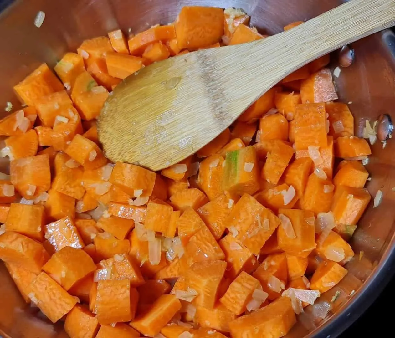 Diced carrots and onions being sautΓ©ed in a pot with a wooden spoon.