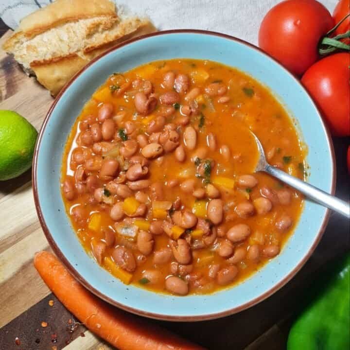 A bowl of easy pinto bean soup with a spoon, surrounded by fresh vegetables and bread.