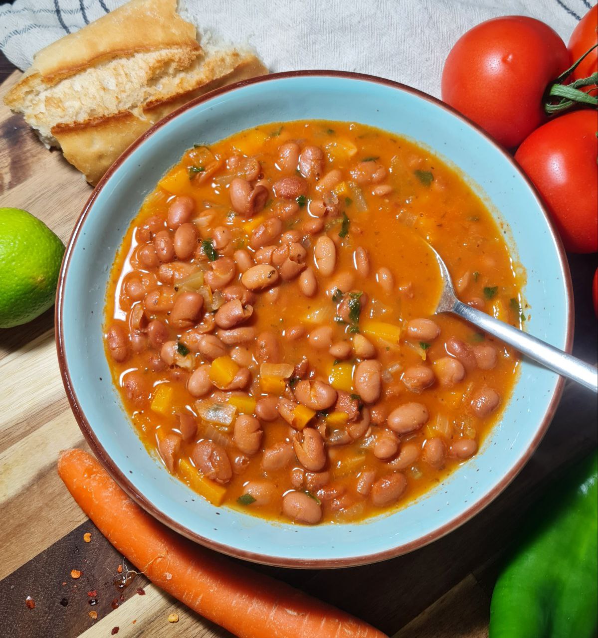 A bowl of easy pinto bean soup with a spoon, surrounded by fresh vegetables and bread.