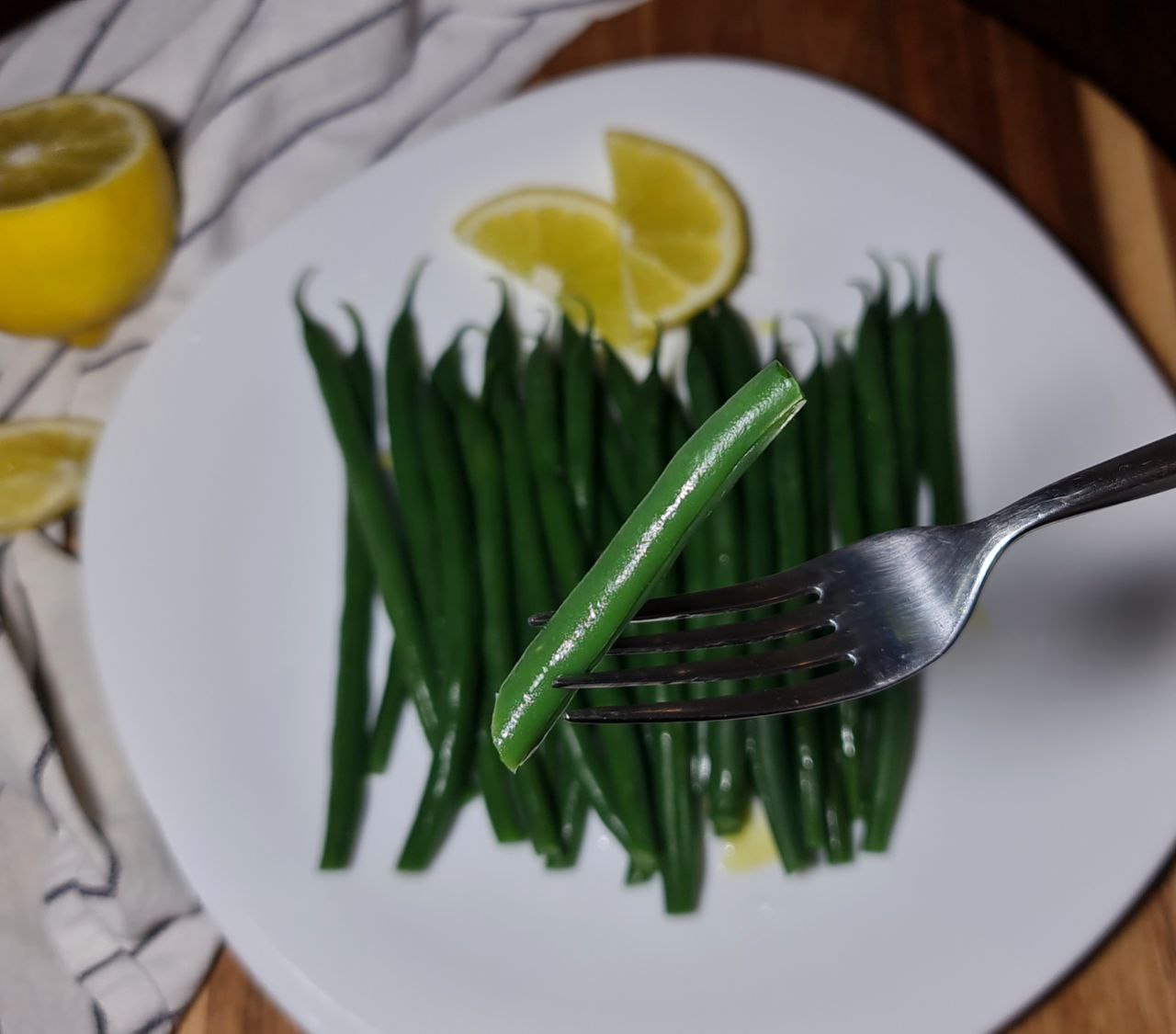 A close-up of boiled green beans on a white plate with lemon wedges, with one green bean held by a fork.