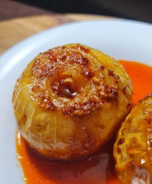 Close-up of a roasted onion with a golden-brown caramelized top, served with a vibrant red sauce on a white plate.