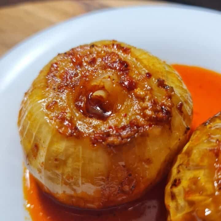 Close-up of a roasted onion with a golden-brown caramelized top, served with a vibrant red sauce on a white plate.