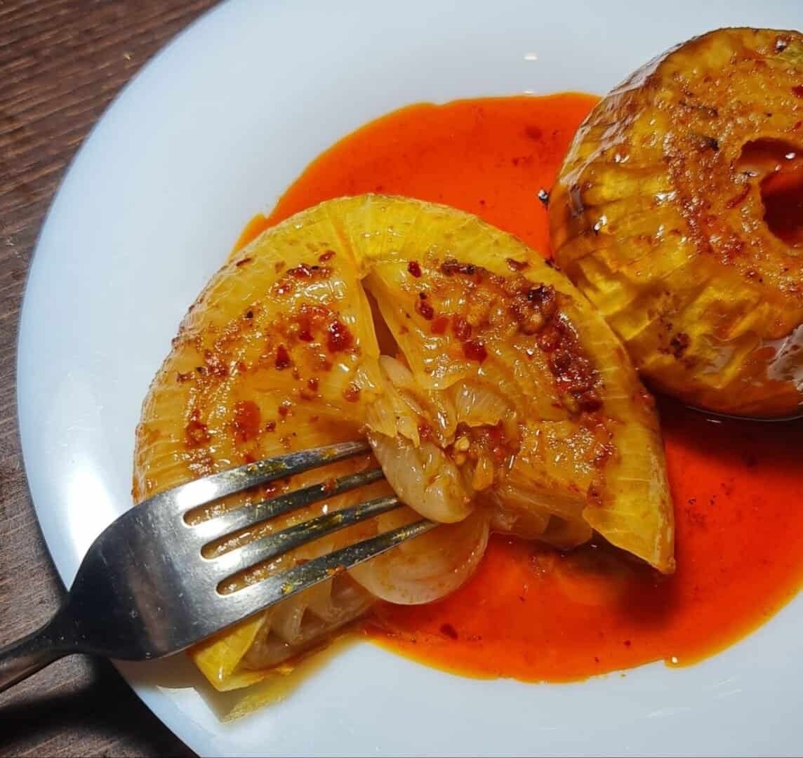 A close-up of a boiled onion dish served on a white plate with a fork, garnished with spices and oil.