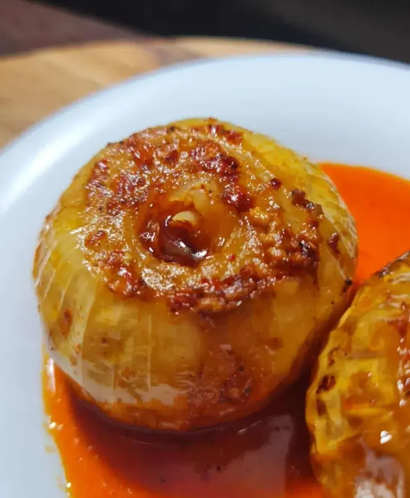 Close-up of a roasted onion with a golden-brown caramelized top, served with a vibrant red sauce on a white plate.