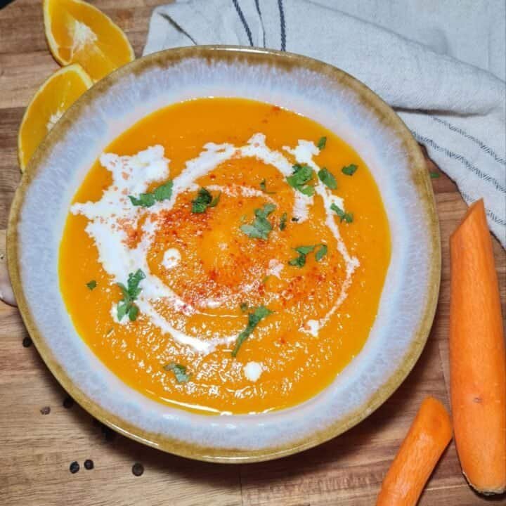 A bowl of vibrant orange carrot soup garnished with a swirl of cream, sprinkled with paprika, and topped with fresh cilantro leaves.