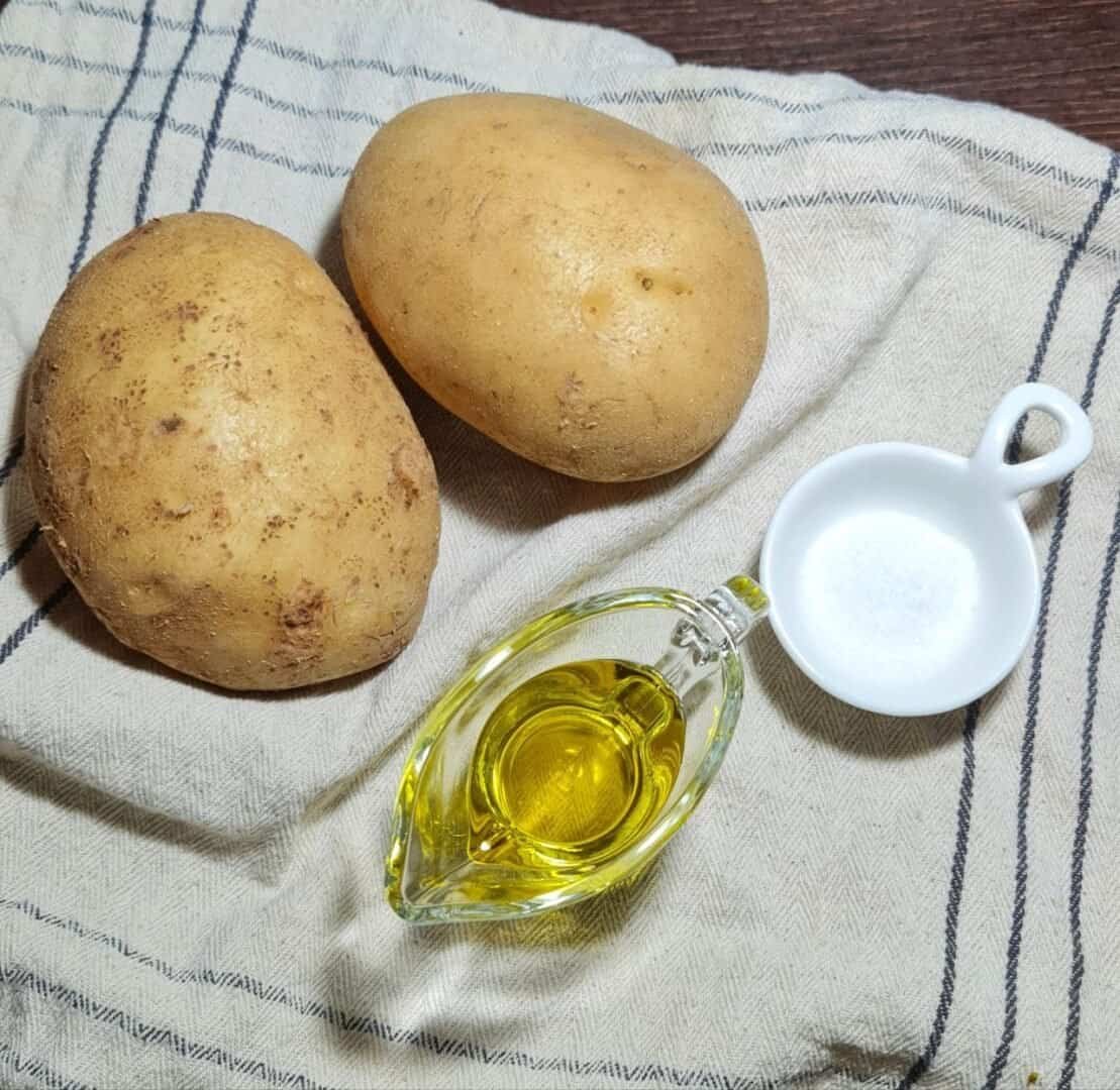 Two raw potatoes, a small glass container of olive oil, and a small white dish of salt on a cloth.